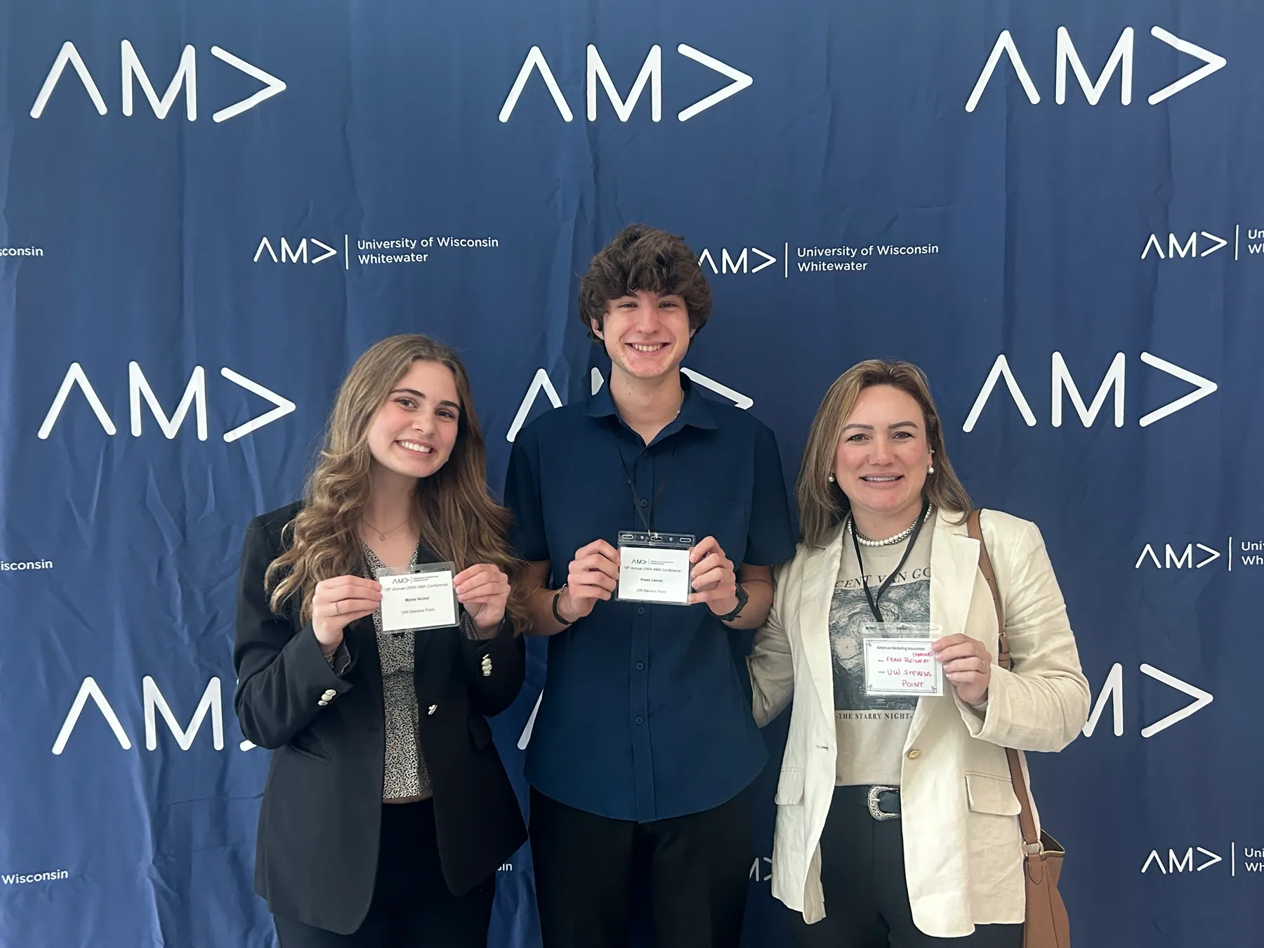 Three attendees holding conference name badges in front of an AMA and University of Wisconsin–Whitewater backdrop.