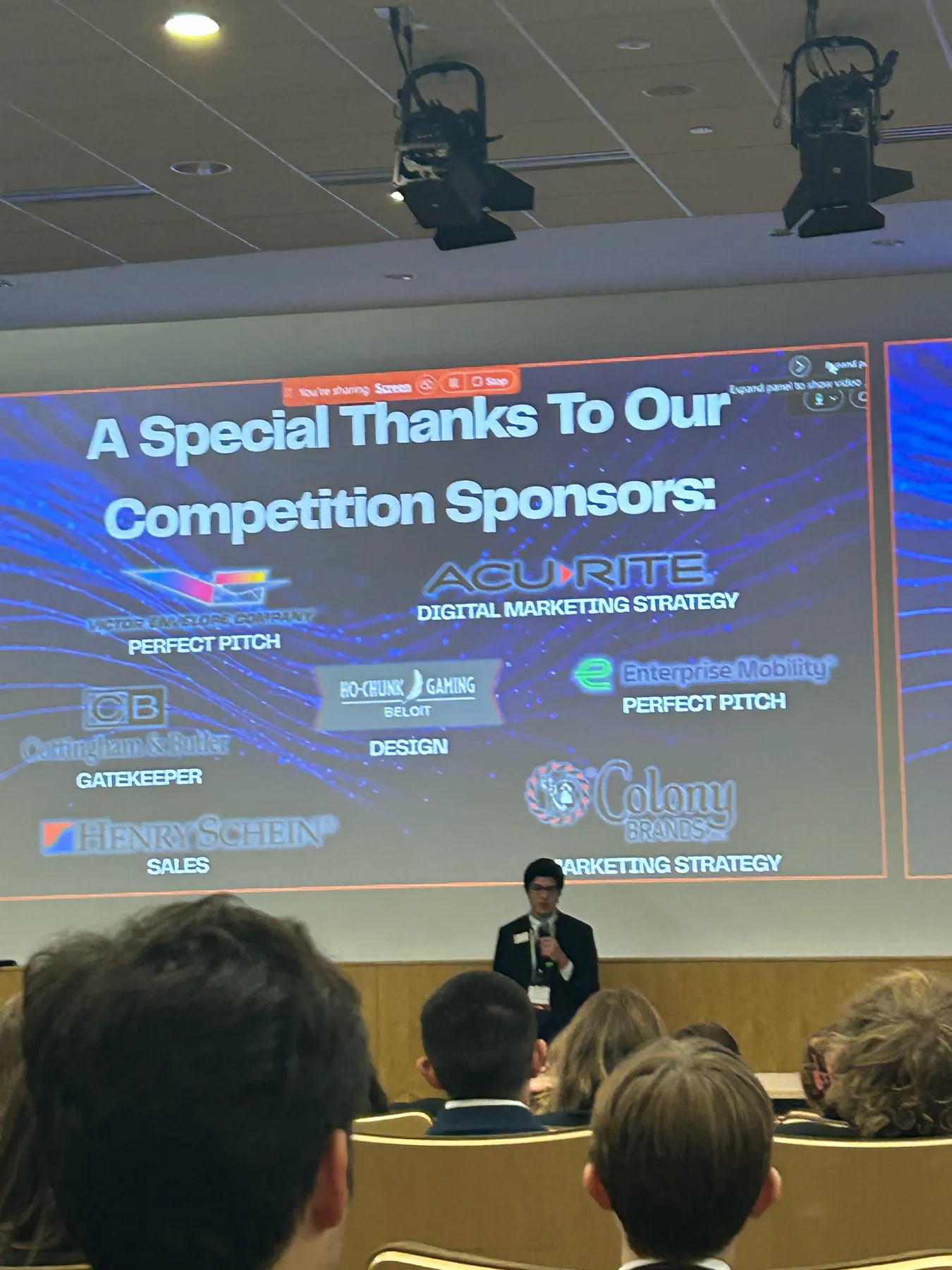 A speaker at the microphone beneath a slide thanking the competition sponsors.