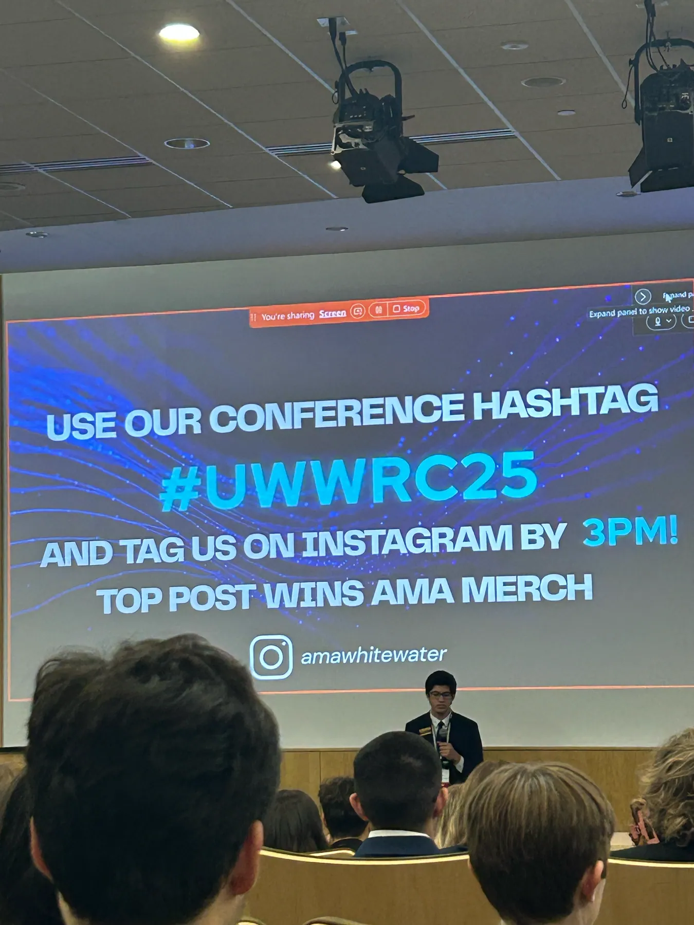 A slide announcing the conference hashtag contest above a speaker at the front of the auditorium.